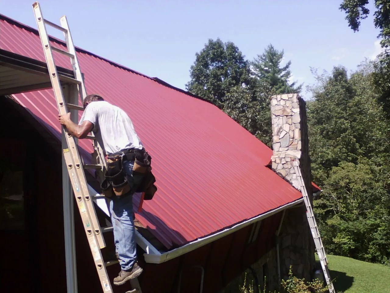 Roofer on a ladder beside a red metal roof and stone chimney — Soffit & Fascia Repair by Roofing Brothers in Fruitland Park